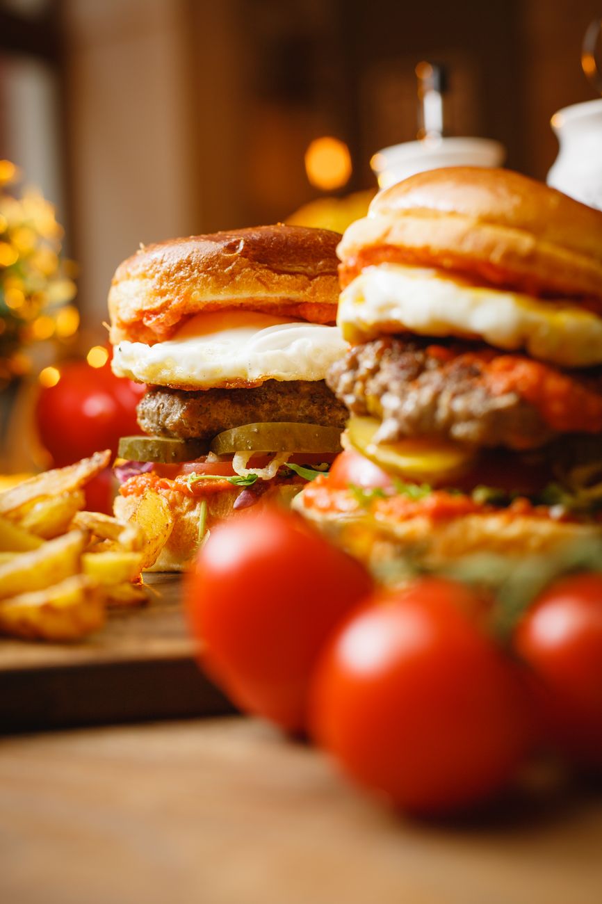 Deux burgers garnis avec oeuf, steak, fromage et cornichons, accompagnes de frites et de tomates, photographies en gros plan dans une ambiance chaude.